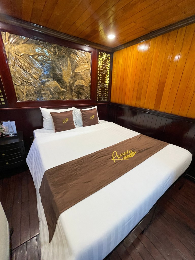 A cozy hotel room featuring a double bed with a brown runner and decorative pillows. The wooden walls and ceiling add warmth, while a carved artwork complements the ambiance.
